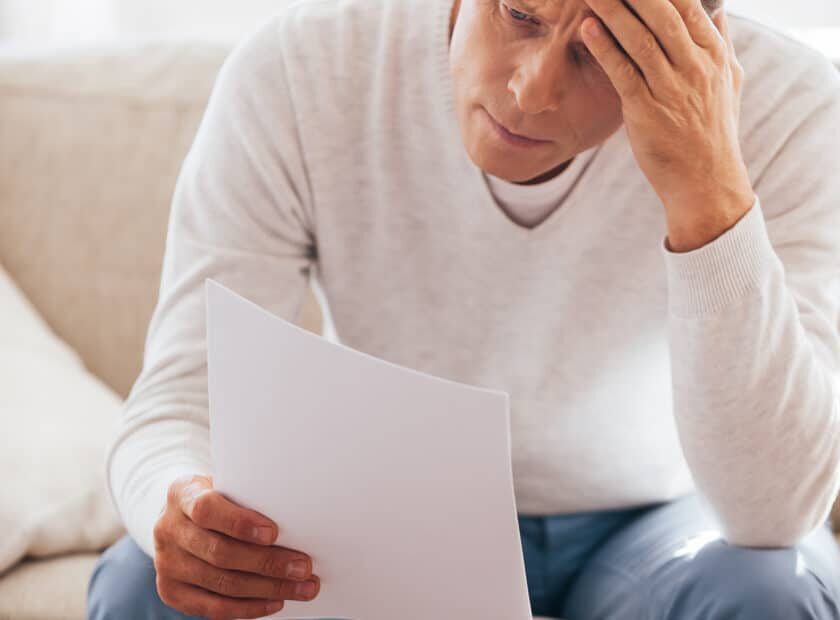 Financial problems. Frustrated mature man holding paper and looking at it while sitting on the couch at home